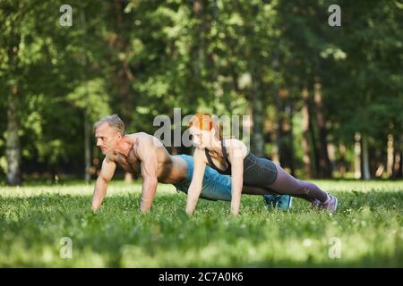 Athletic mature couple doing push-ups on grass while training together in park Stock Photo
