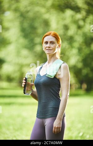 Portrait of smiling confident redhead mature woman with towel on shoulder holding bottle of water in summer park Stock Photo