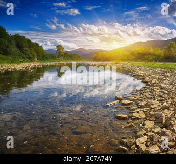 Mountain river stream of water in the rocks with blue sky. Clear river with rocks leads towards mountains  lit by sunset Stock Photo