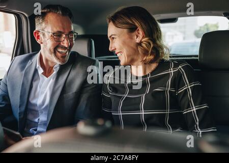 Two business people traveling in backseat of a car. Businessman and businesswoman looking at each other and smiling while riding a taxi to work. Stock Photo