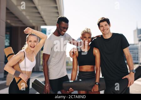 Multi-ethnic friends with fitness mats standing outdoors after workout. Fitness group smiling after workout. Stock Photo