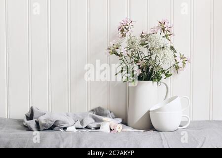 Bouquet of pink soapwort and wild carrot flowers in ceramic pitcher. Empty coffee cups on linen table cloth. White wooden wall background. Feminine Stock Photo