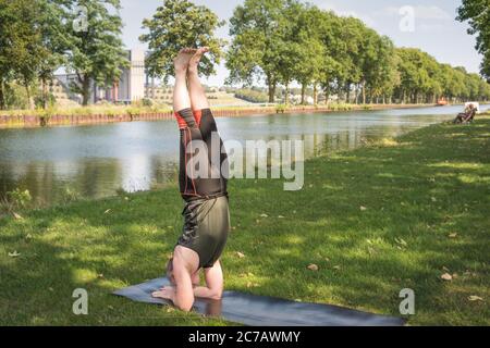 A man does an upside down yoga pose on the grass by a channel with a fisher at the background. Stock Photo