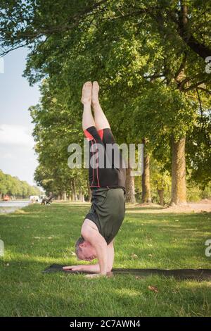 Strong man does a hard twisted upside down yoga pose, a variation of supported headstand or Sirsasana, outdoors in a summer day. Stock Photo