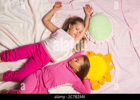 Kids in pink pajamas lie close and look at each other. Children with sleepy faces on light pink blanket background. Schoolgirls have pajama party with funny pillows. Childhood and happiness concept Stock Photo