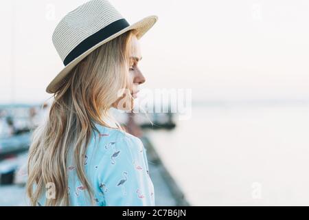 Beautiful blonde young woman in blue dress and straw hat on pier on sunset