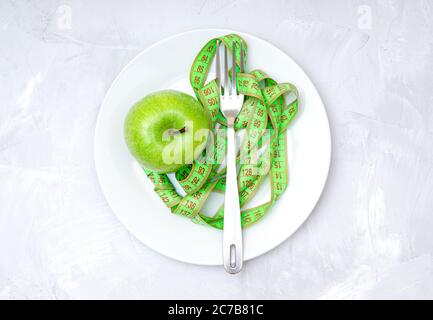Soft measuring tape, fresh green apple and a fork on a white plate on a concrete table, top view. Weight loss fruit diet concept. Stock Photo