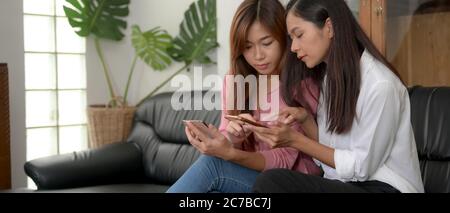 Side view of two female teenagers using smartphone and talking to each other while sitting in living room Stock Photo