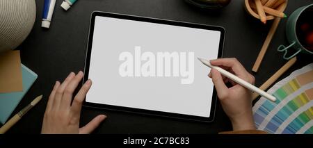 Overhead shot of female designer drawing on mock up tablet with stylus pen on dark worktable with painting tools Stock Photo