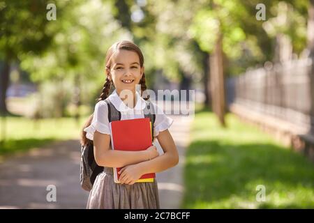 Portrait of happy little girl with backpack standing on city street. Cheerful child in uniform on her way to school Stock Photo