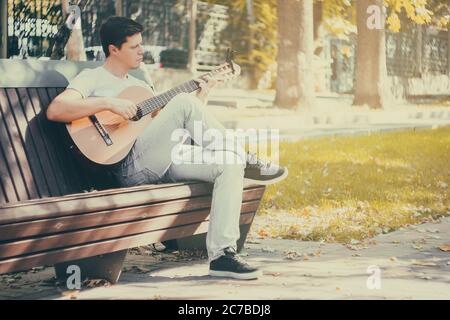 Young man sitting on the bench in the park playing on acoustic guitar Stock Photo