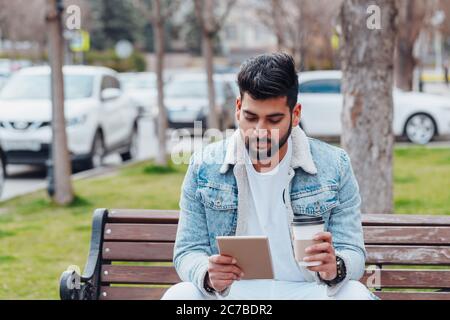 A bearded stylishly dressed Indian man sits on a bench and uses his tablet. Stock Photo