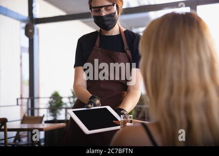 The waitress works in a restaurant in a medical mask, gloves during coronavirus pandemic. Representing new normal of service and safety. Getting paid contactless, taking care of clients. Stock Photo