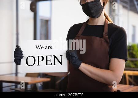 The waitress works in a restaurant in a medical mask, gloves during coronavirus pandemic. Representing new normal of service and safety. Holding board with words WE ARE OPEN. Keeps distance. Stock Photo