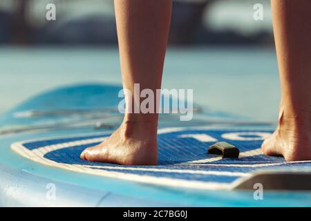 Close up legs. Young attractive woman standing on paddle board, SUP. Active life, sport, leisure activity concept. Caucasian woman on travel board in summers evening time. Vacation, resort, enjoyment. Stock Photo