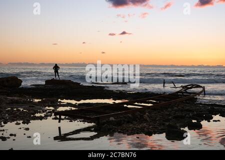Silhouette of woman looking away at the Mediterranean sea, near an abandoned metal pier on a beach, at sunset,  in Batroun, Lebanon, Middel East Stock Photo