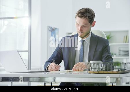 Concentrated handsome young businessman in gray suit sitting at desk and planning tasks while working in office Stock Photo