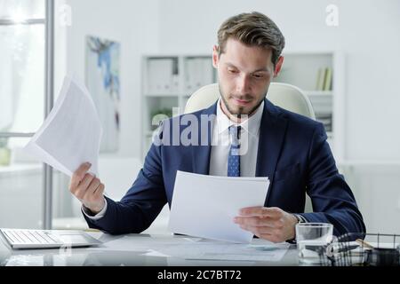 Busy young manager in formal suit sitting at desk and examining business papers in office Stock Photo