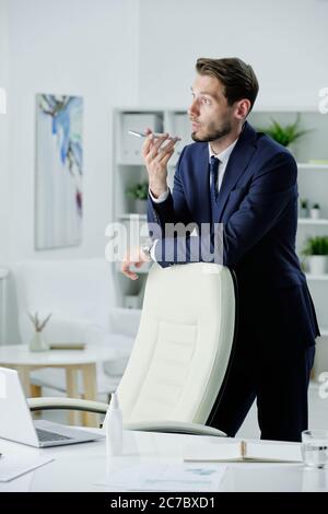 Confident young businessman in formal suit leaning on office chair while sending voice message to colleague Stock Photo