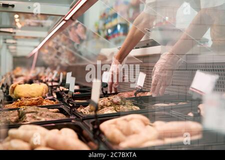 Close up of raw meat pieces in refrigerator with price tags ready for sale in meat department of store. Male hands putting out of glass counter plate with sliced fresh steaks. Concept of food. Stock Photo