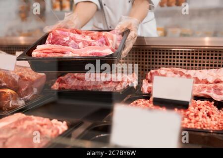 Close up of raw meat pieces in refrigerator with price tags ready for sale in meat department of store. Female hands putting out of glass counter plate with sliced fresh steaks. Concept of food. Stock Photo