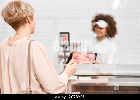 Young smiling woman with curly hair demonstrating to lady raw pork. Cheerful girl behind store counter with big assortment showing meat in bowl from refrigerator, supermarket. Stock Photo