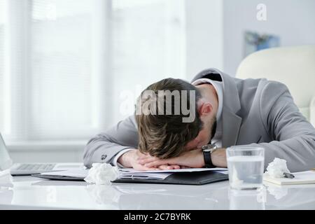 Exhausted young businessman lying on table with papers and crumpled napkins while suffering from headache Stock Photo