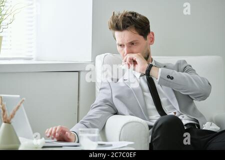 Frowning ill young businessman sitting in armchair and working on project using laptop while having cold Stock Photo