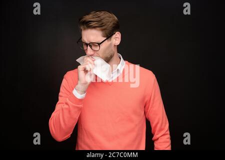 Sick young man in eyeglasses standing against black background and sneezing into napkin, virus concept Stock Photo