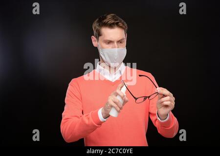 Focused young man in cloth mask cleaning eyeglasses with disinfectant, coronavirus prevention concept Stock Photo