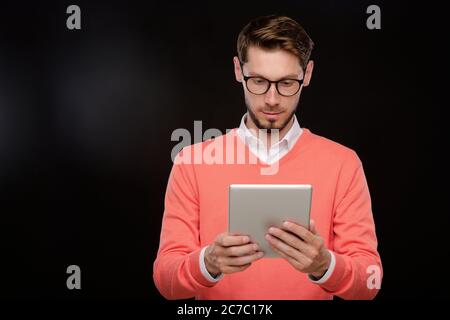 Concentrated young bearded man in orange sweater using modern tablet against black background, social media concept Stock Photo