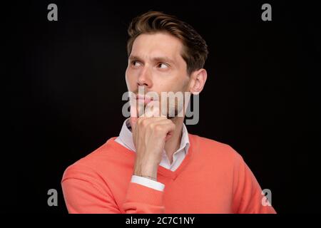 Pensive handsome young man in orange sweater rubbing chin while thinking of strategy, black background Stock Photo