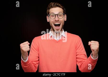 Portrait of excited young bearded man with open mouth making yes gesture while winning prize, black background Stock Photo