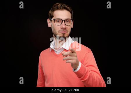 Portrait of young bearded man in orange pleasantly surprised pointing with finger at camera against black background Stock Photo