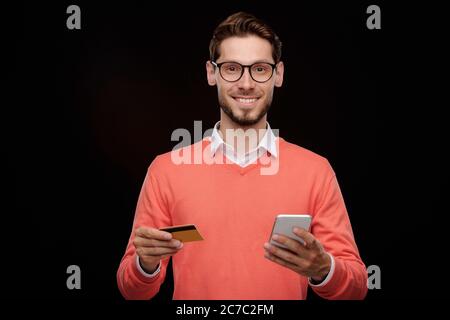Portrait of cheerful handsome young man with stubble standing with credit card and smartphone and using online transaction service Stock Photo