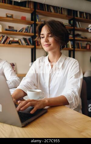 Young beautiful smiling woman sitting at the table while happily working on laptop with cup of coffee in office Stock Photo