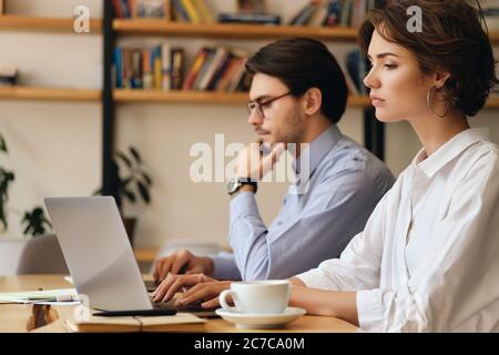 Young tired woman sitting at the table thoughtfully working on laptop with colleague near in office Stock Photo