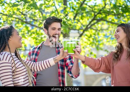 Guy and two girls touching glasses of coffee standing under tree Stock Photo