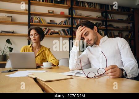 Young tired man sitting at the table with notepad and eyeglasses while thoughtfully holding hand near head at work in office with colleague on Stock Photo