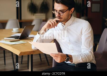 Young serious businessman in eyeglasses thoughtfully reading papers while working in modern office Stock Photo