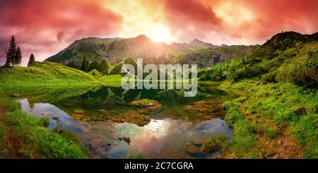 Dramatic sunrise at a lake in the Alps with mountains, red clouds reflected in the clear water and paths leading through the vibrant green grass Stock Photo