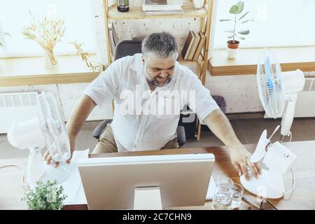 Extremely. Businessman, manager in office with computer and fan cooling off, feeling hot, flushed. Using fan but still suffering of uncomfortable climate in cabinet. Summer, office working, business. Stock Photo