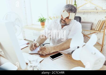 Can't concentrate. Businessman, manager in office with computer and fan cooling off, feeling hot. Using fan but still suffering of uncomfortable climate in cabinet. Summer, office working, business. Stock Photo