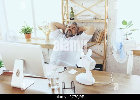 Tired off. Businessman, manager in office with computer and fan cooling off, feeling hot, flushed. Using fan but still suffering of uncomfortable climate in cabinet. Summer, office working, business. Stock Photo