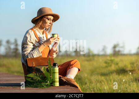 Woman botanist with backpack sitting on wooden path through peat bog swamp in wildlife national park. Naturalist resting on boardwalk, drinking tea, e Stock Photo