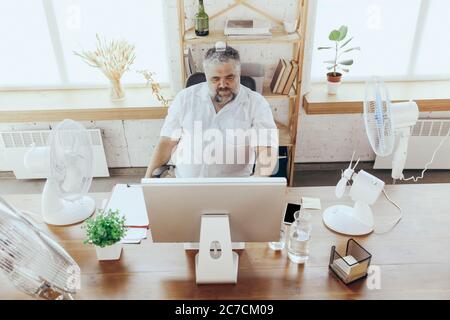 Ice helping. Businessman, manager in office with computer and fan cooling off, feeling hot, flushed. Using fan but still suffering of uncomfortable climate in cabinet. Summer, office working, business. Stock Photo
