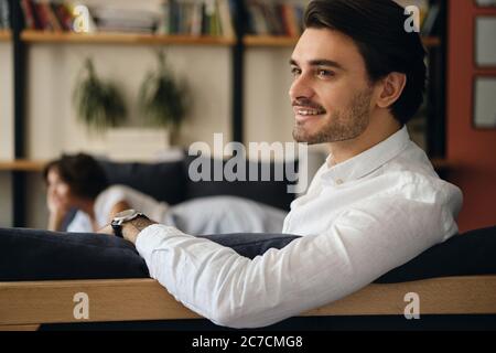 Young attractive smiling businessman sitting on sofa with colleague while happily looking aside in modern co-working space Stock Photo