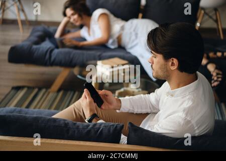 Side view of young businessman sitting on sofa with colleague on background while thoughtfully using cellphone in co-working space Stock Photo