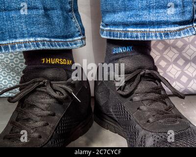 Man sitting down on a bed quilt wearing denim jeans, with feet in casual shoes wearing odd socks – different days of the week sewn into the fabric. Stock Photo