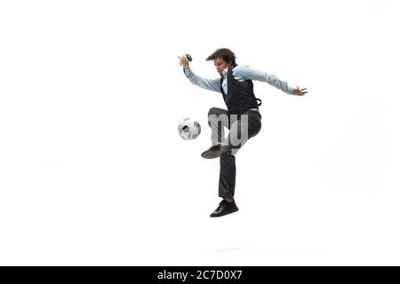 Man in office clothes playing football or soccer with ball on white background like professional player. Unusual look for businessman in jump kicking ball. Sport, healthy lifestyle, creativity. Stock Photo